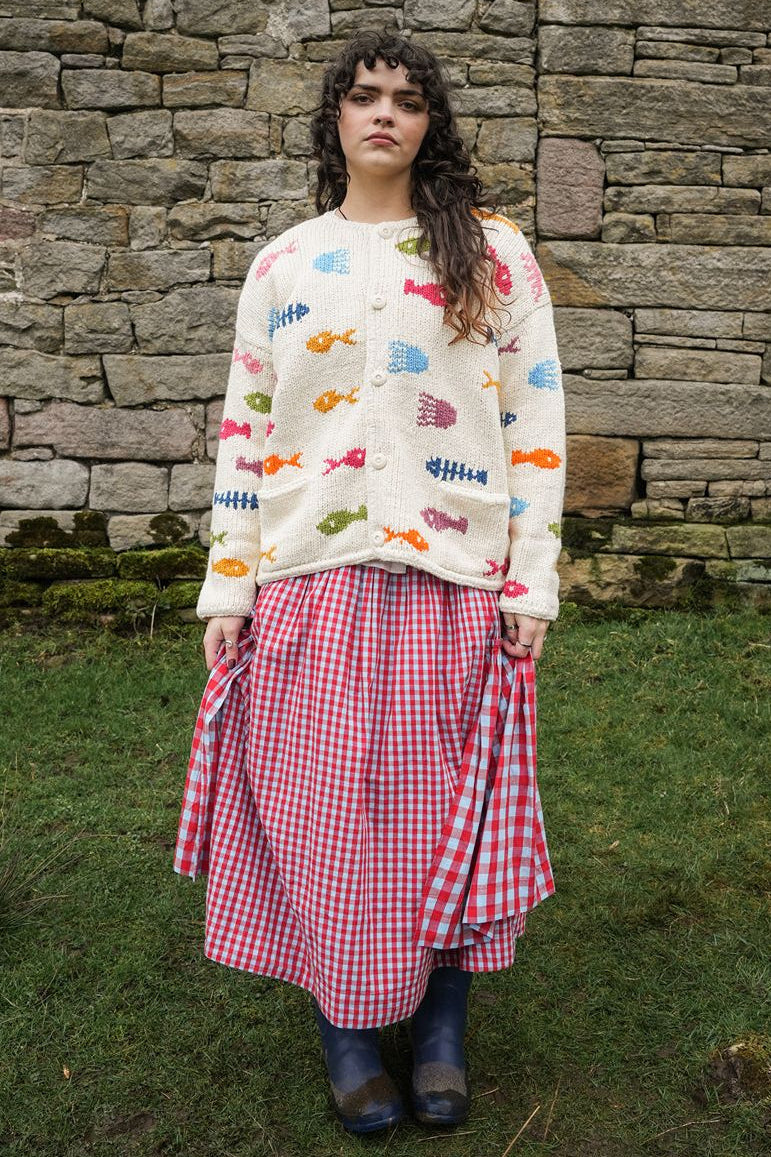 Person wearing a colorful sweater and red checkered skirt standing in front of a stone wall.